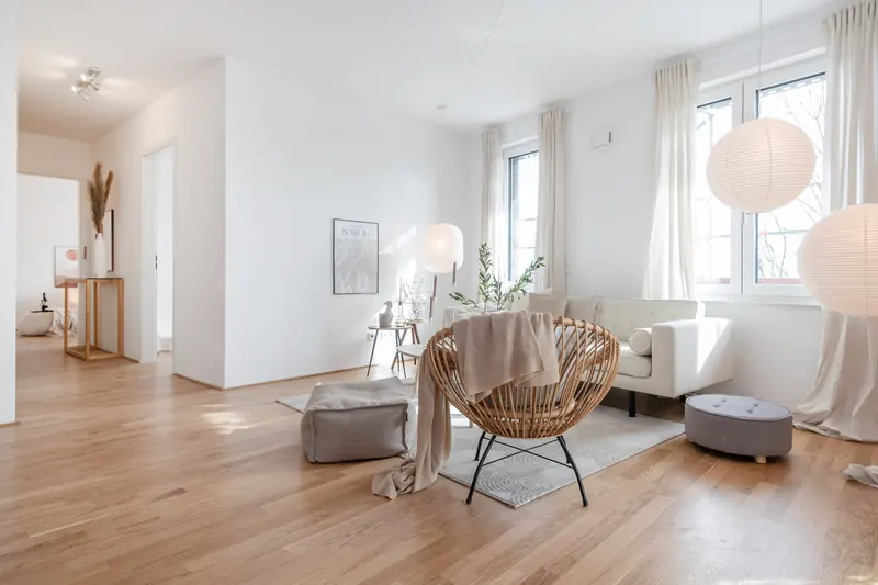 Wohnbereich mit Rattansessel, Sitzpoufs und hellen Vorhängen vor großen Fenstern