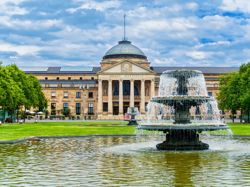 Kurhaus Wiesbaden mit Säulenportal und Kuppel hinter dem Bowling Green mit Springbrunnen unter hellblauem Himmel